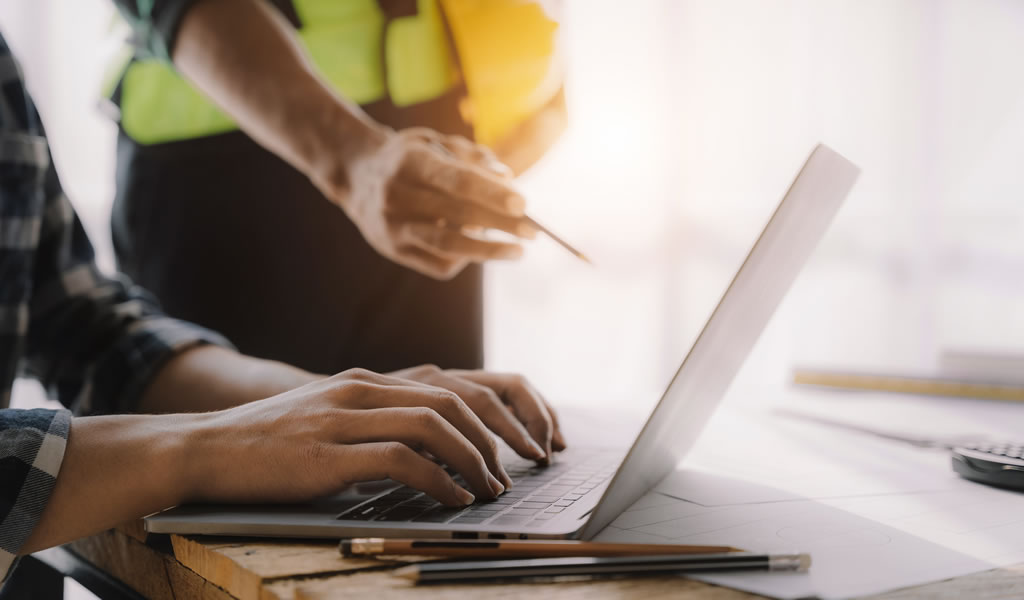 Hand pointing, hands working on laptop at construction site at desk in office.